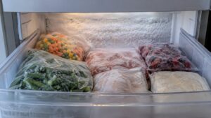 The inside of an open freezer drawer with a layer of white frost on the back wall and frozen packages